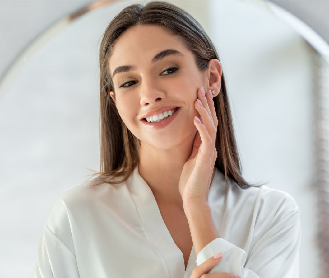 A woman with long brown hair wearing a white shirt smiles and touches her face.